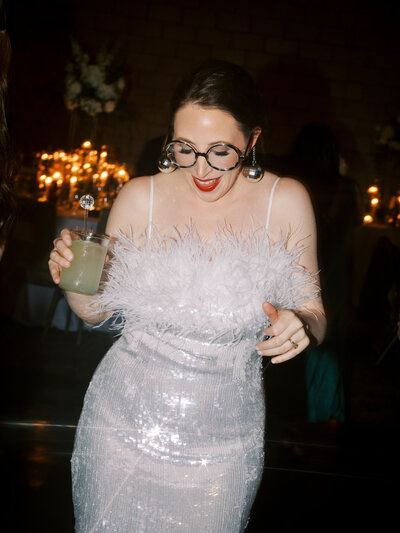 bride in feathered sequin dress dancing with a drink in her hand and disco ball glasses