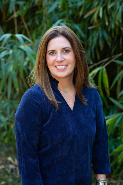 Woman with long brown hair, wearing a blue top, smiling and standing in front of a backdrop of green foliage.