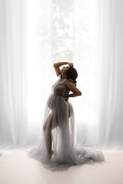 woman with black hair wearing a sheer gray tulle gown in front of windows with white drapes