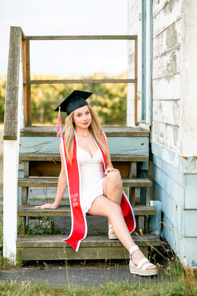 A high school senior sits on a set of wood stairs in a white dress and her cap and stole
