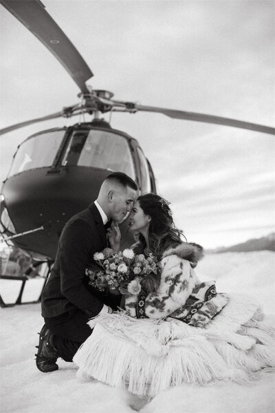 couple sitting forehead to forehead in front of helicopter on a glacier