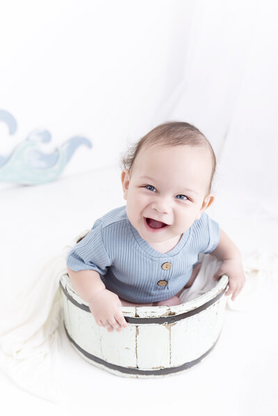 A toddler boy sits in a wooden bucket in a blue onesie while smiling big