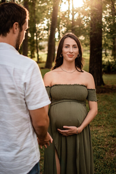 An expectant mother stands in a field of pine tress and holds her belly and her husband's hand.