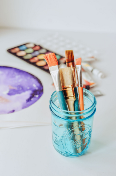 Paintbrushes in an aqua glass jar with a watercolor paint palette in the background - Bloom by bel monili