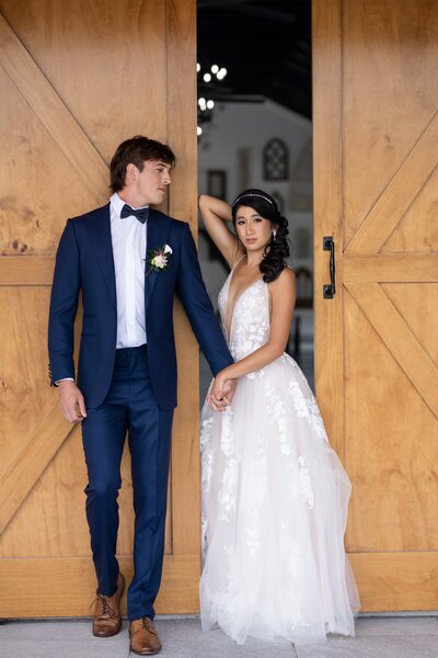 Couple leans against a barn door as the bride looks at the camera and groom gazes at the bride