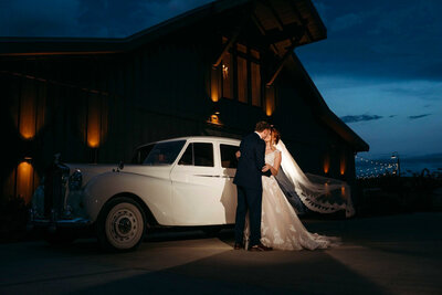 But a bride and groom kissing in front of a vintage car with  an illuminated barn in the background at dusk