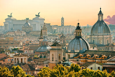 A panoramic view of a cityscape featuring a cluster of historic buildings with domes and spires. In the distance, a large monumental building with statues on top is visible. The sky is warm, suggesting sunset or sunrise.