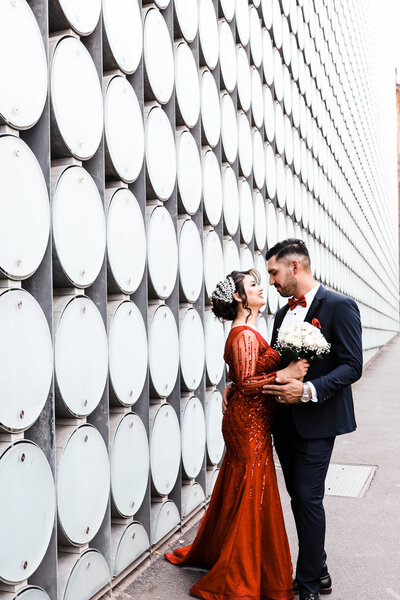 bride wearing a red gown holding flowers while the groom holds her and look at each other as they stand beside the wall with round glass structures