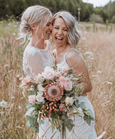 two brides with bouquet in grass field, hugging and laughing