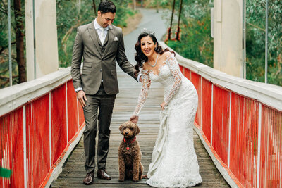 a caboodle dog sitting between a bride and groom  while the stand on a red bridge