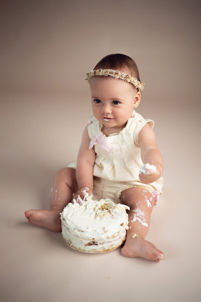 One year old toddler wearing an ivory romper and floral halo crown celebrating her first birthday milestone with a cake smash photo session in Collingwood Ontario