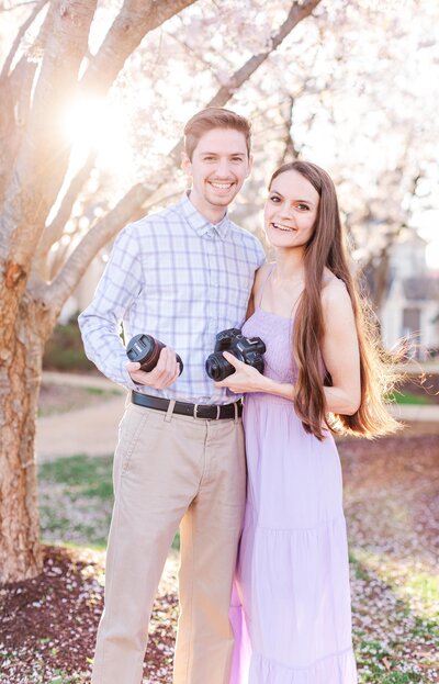 Photographers wearing lavender dress and blue button up shirt, smiling while holding cameras in front of cherry blossom trees during spring in Nashville