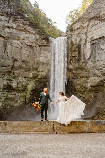 Bride holds out wedding dress holding hands with groom while standing in front of Taughannock Falls in upstate new york.