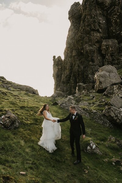 A bride and groom hold hands, walking on grassy hills with large rocky cliffs in the background—capturing real moments during their Colorado wedding day.