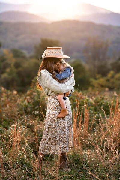 A mom and daughter snuggle in the tall grass below a mountain ridge
