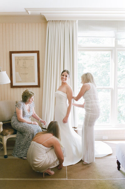 Bride smiling at camera while three women help her dress