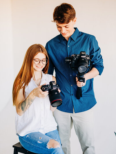 This image is of a husband and wife photography and videography duo. The woman is wearing a black dress and a camera harness, and is holding one camera up as she prepares to take a photo. The man is wearing a grey polo and black dress pants, and holds a gimbal with a camera attached.