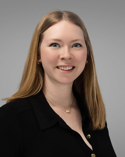A person stands on stone steps in front of a large stone building, holding a bright red coat. They are wearing a white dress and red shoes. Trees are visible in the background.