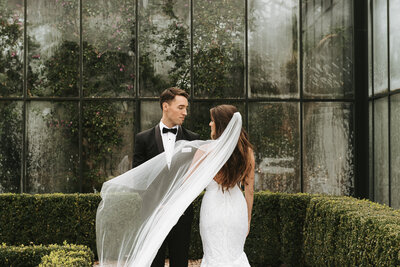 Bride and groom posed on wedding day in front of a botanical garden with a veil