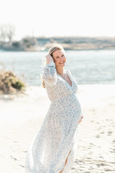 A pregnant woman wearing a whtie dress with blue flowers stands on the beach holding her pregnant belly and brushing her hair out of her face photographed by New jersey maternity photographer Kate Voda Photography