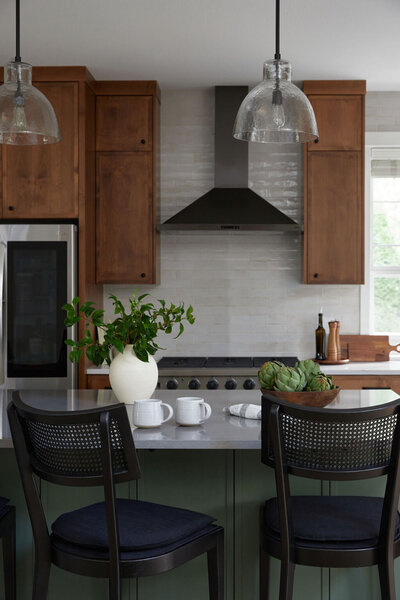 Kitchen with medium brown cabinets and beige subway tile backsplash with a gas range and silver range hood. Green island in the front with Three black barstools with cane backrests. A white vase of greenery, a bowl of artichokes, and two white mugs sit on the island.
