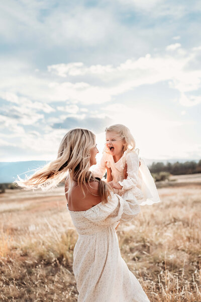 mom in white dress lifting her blonde daughter in the air and they're both smiling