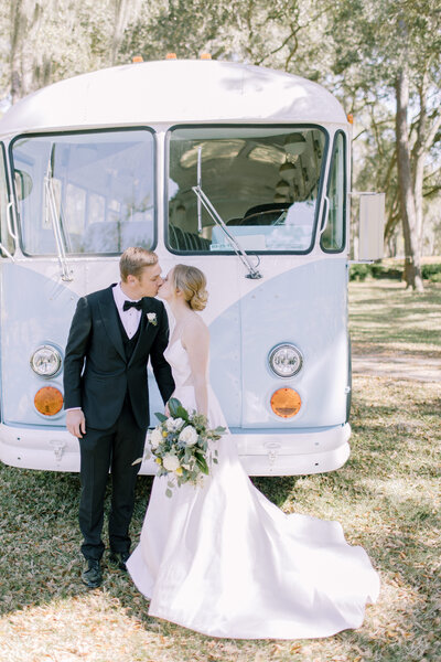 This savannah wedding photo is of emily in white spaghetti strap gown and ross in black tux and bowtie standing in front of a vintage, baby blue bus under the shade of many oak trees draped with spanish moss.
