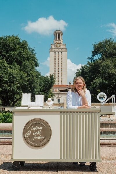 Owner of Lasting Links Austin standing behind the jewelry cart, featuring elegant displays of permanent bracelets and necklaces in front of the UT Tower.