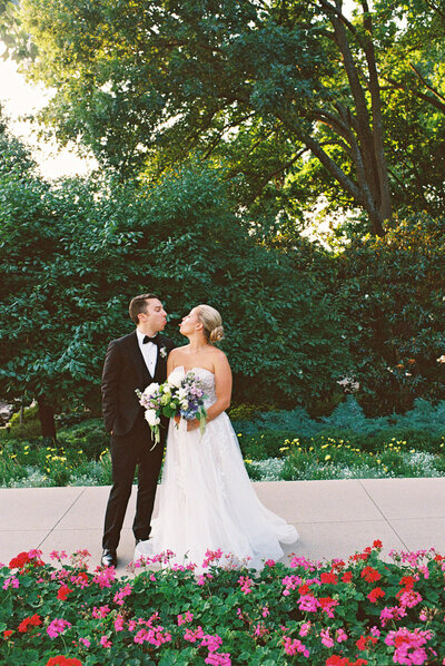 A newly married couple stands before a lush green backdrop, sharing a playful, intimate moment, their joy captured amidst vibrant flowers