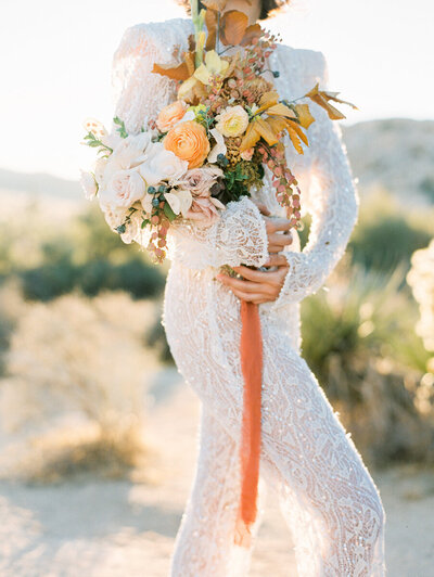 Bride stands in joshua tree desert wearing beaded long sleeve gown and oversize wedding bouquet with orange flowers at Joshua Tree National Park