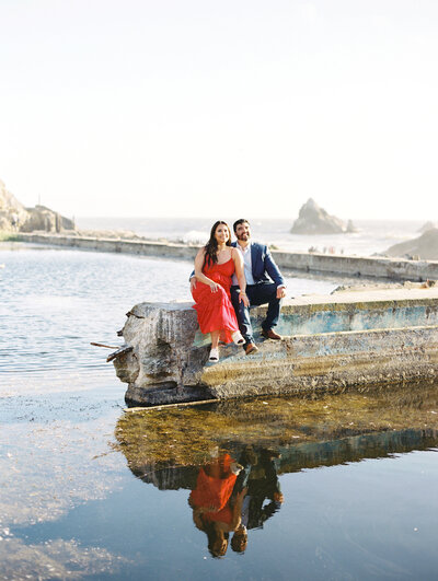 Couple sitting on sutro baths ruins in San Francisco with girl wearing red dress and guy wearing a suit, photo by Anastasiya Photography - San Francisco Photographer