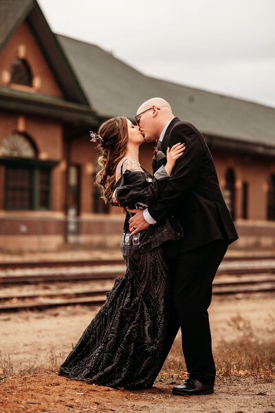 A newlywed couple kissing in front of a historic train station in downtown Bemidji, Minnesota. The woman is wearing a black dress, black denim jacket with patches, and autumnal florals. Captured by Alyssa Daken Photography.