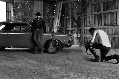 Country Music Photography behind the scenes Mark Maryanovich kneeling while looking at back of camera Chance Moore standing next to vintage car inside barn with lights hanging from ceiling