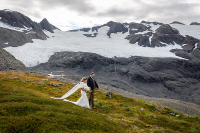 Bride and groom walk down a grassy hill while a helicopter rests behind them.