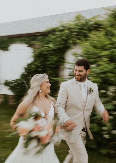 Motion blurred photo of bride with pink, white and green bouquet holding hands and running with husband in tan suit in front of ivy covered wedding venue