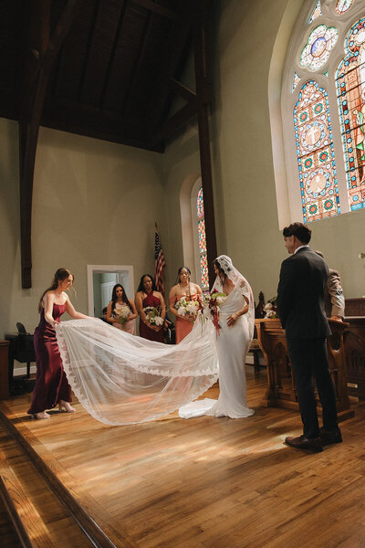 bridesmaid fluffing the brides veil illuminated by the sun coming through a stained glass window in pensacola florida