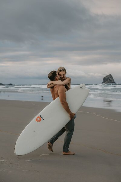 Man carrying woman and surfboard on a sandy beach with waves and a cloudy sky in the background, capturing the spirit of an Oregon coast photography retreat for wild-hearted artists.