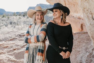 A Mom and daghter in cowboy hats smile together outdoors by rocky cliffs and desert scenery during a playful family photo session in Montrose, Colorado.