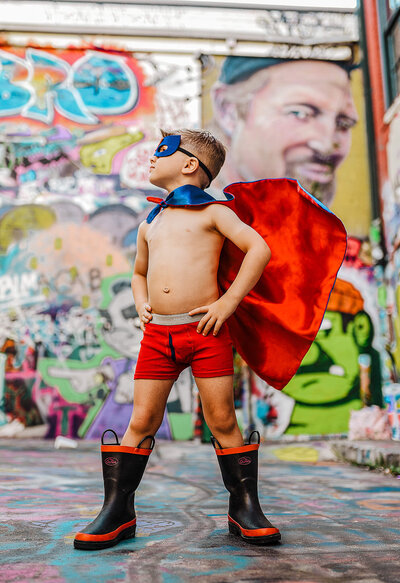 Boy in underwear with rainboots and superhero cape and mask posing in graffiti alley in baltimore Maryland near  MICA