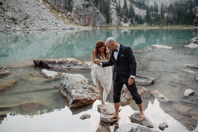 Delta Lake wedding with groom helping his bride through the lake on  rocks for their adventure elopement with jackson hole photographers