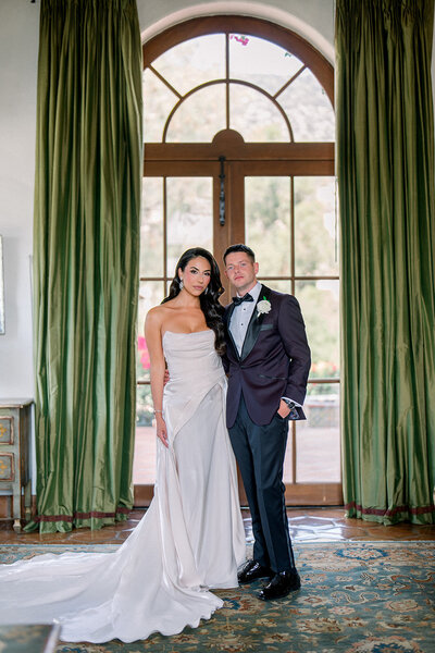 Bride and groom pose in front of tall arched window framed by velvet green curtains at Hummingbird Nest Ranch, captured in timeless editorial wedding photography.