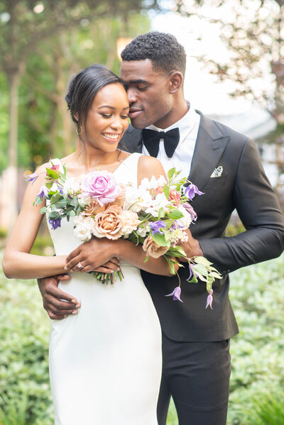 Groom in tuxedo embraces his bride while she holds a bouquet