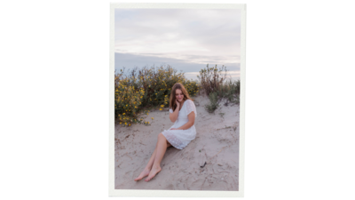 Senior girl sits on dune in Galveston Beach while wearing a white dress