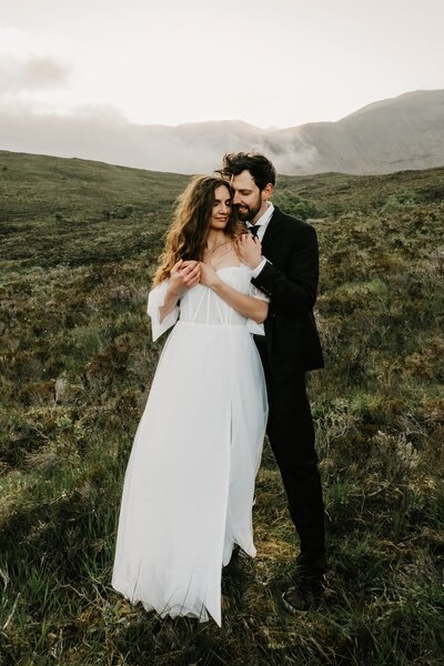 Bride in white dress and groom in black suit embrace outdoors on a grassy hillside, capturing stunning wedding portraits with a Colorado mountain backdrop.