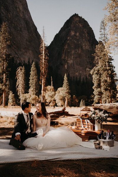 a bride and groom sitting on the ground, having a picnic with a mountain/forest view