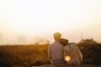 A couple sits on the edge of a cliff, the woman has her hand on the mans shoulder, they are watching a sunset