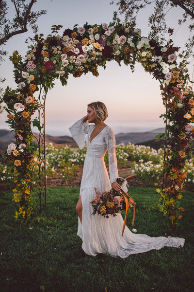 Boho bride standing under a fall floral ceremony arch holding a bouquet