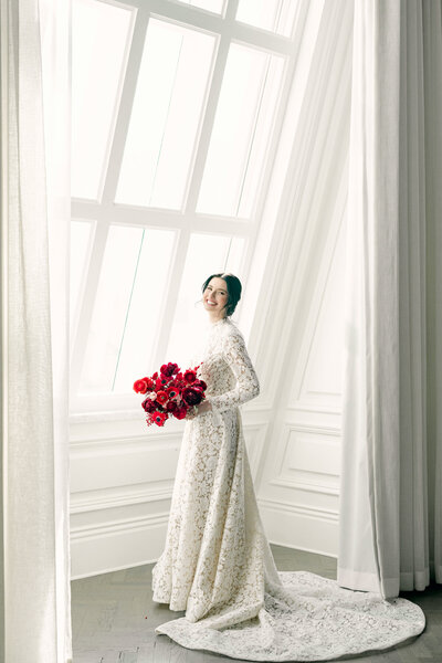 Bridal portrait of bride standing by window holding bouquet of red flowers at luxury wedding in Dallas