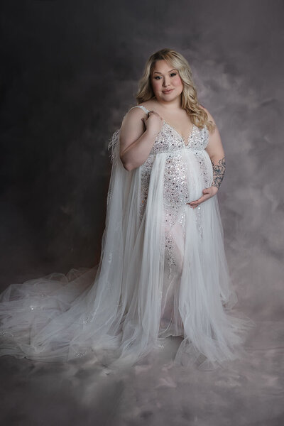 A mother to be stands in the studio of a New Orleans Family, Maternity & Newborn Photographer holding her bump in a white maternity gown