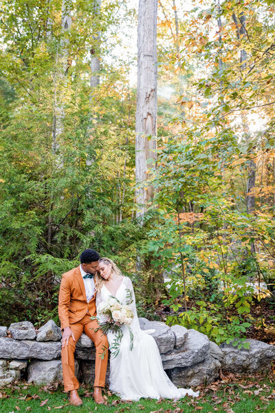 A groom and bride sit on a stone bench. He is wearing an orange suit and is kissing her on her forehead.